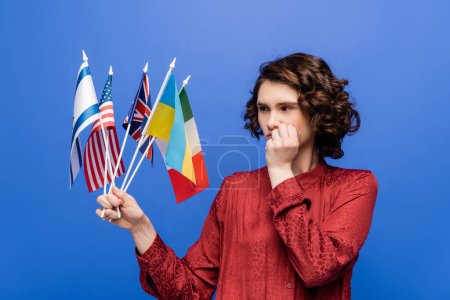 nachdenkliche Frau, die die Hand vor dem Gesicht hält, während sie Flaggen verschiedener Länder betrachtet, die auf blauem Grund isoliert sind