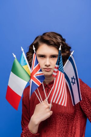 estudiante joven y seguro de sí mismo con diferentes banderas internacionales mirando a la cámara aislada en azul