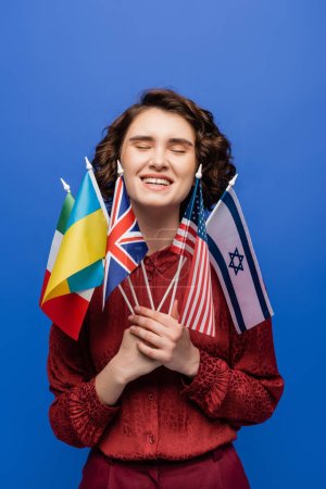 joven feliz sosteniendo banderas internacionales y sonriendo con los ojos cerrados aislados en azul