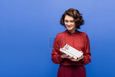 zufriedene Studentin in roter Bluse beim Anblick fremdsprachiger Lehrbücher isoliert auf blauem Grund