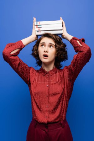 shocked student opening mouth and holding textbooks of foreign languages over head isolated on blue