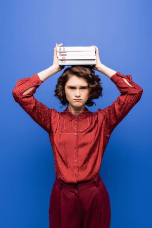 displeased student holding textbooks of foreign languages above head and looking at camera isolated on blue