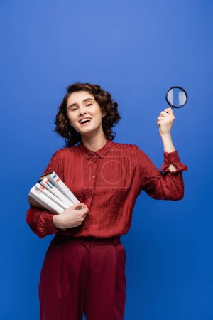 carefree student with magnifier and textbooks of foreign languages smiling at camera isolated on blue