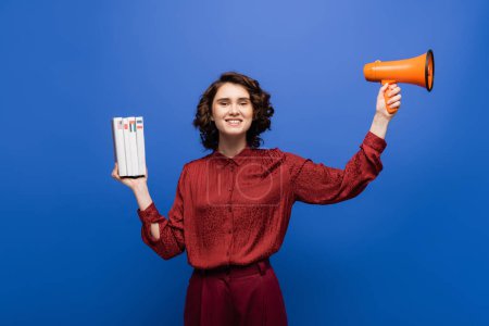excited language teacher holding megaphone and textbooks while smiling at camera isolated on blue