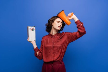 exhausted language teacher standing with closed eyes while holding megaphone and textbooks isolated on blue