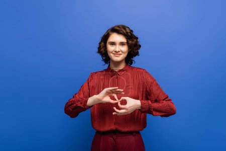 Téléchargez les photos : Jeune femme joyeuse regardant la caméra et montrant le symbole signifiant interprète sur le langage des signes isolé sur bleu - en image libre de droit