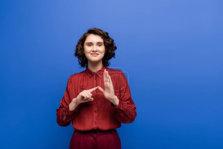 happy young teacher showing alphabet on sign language and looking at camera isolated on blue