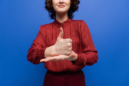 partial view of positive woman in burgundy blouse showing gesture meaning help on sign language isolated on blue