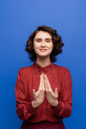 positive teacher smiling at camera and showing gesture meaning happy on sign language isolated on blue