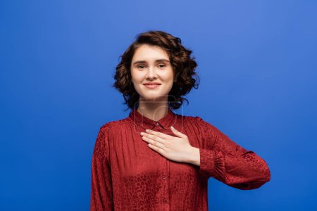 cheerful woman looking at camera and telling please on sign language isolated on blue