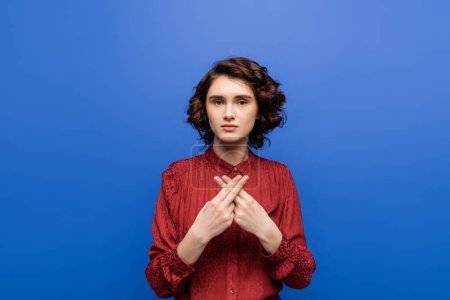 young brunette woman looking at camera while using sign language isolated on blue