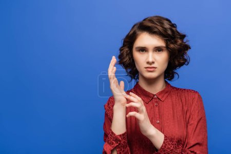 young woman looking at camera while gesturing using sign language isolated on blue