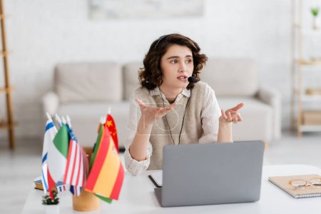 brunette language teacher in headset gesturing near laptop and blurred international flags at home