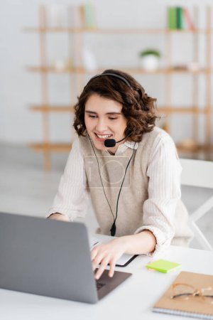 junge und glückliche Sprachlehrerin tippt im Headset auf Laptop, während sie zu Hause Online-Unterricht hat