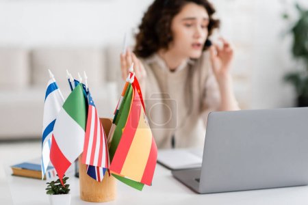 enfoque selectivo de diferentes banderas internacionales cerca de la computadora portátil y difuminado profesor de idiomas que trabaja en casa