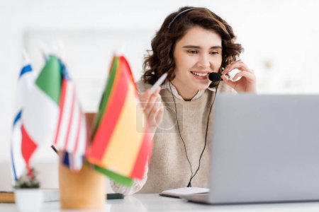 Photo for Overjoyed language teacher in headset holding pen and talking near laptop and international flags on blurred foreground - Royalty Free Image