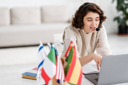 profesor de idiomas positivo en auriculares hablando durante la lección en línea en el portátil cerca de banderas internacionales en casa