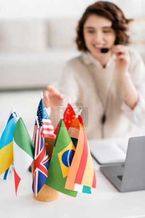 Selektiver Fokus verschiedener internationaler Flaggen in der Nähe von Laptop und unscharfem Sprachlehrer, der zu Hause arbeitet