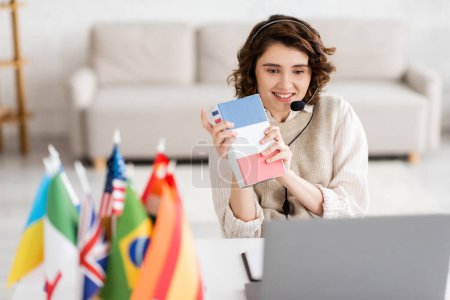 Photo for Smiling language teacher in headset showing French textbook during video lesson on laptop near blurred international flags - Royalty Free Image