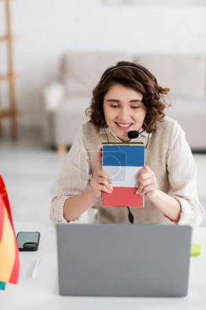 profesor alegre en auriculares que muestra el libro de texto de la lengua francesa durante la lección en línea en el ordenador portátil en casa