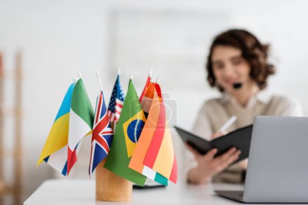 Selektiver Fokus internationaler Flaggen in der Nähe von Sprachlehrern, die während des Online-Unterrichts zu Hause in ein Notizbuch schreiben