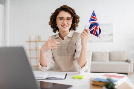 young language teacher in eyeglasses holding flag of United Kingdom near laptop on blurred foreground at home