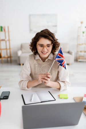 cheerful language teacher in eyeglasses holding flag of United Kingdom while working near notebook and laptop at home