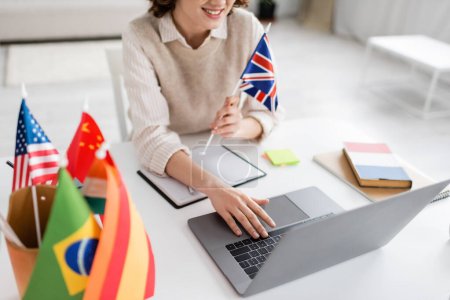 Téléchargez les photos : Vue recadrée du professeur de langue souriant avec drapeau du Royaume-Uni en utilisant un ordinateur portable pendant la leçon en ligne à la maison - en image libre de droit
