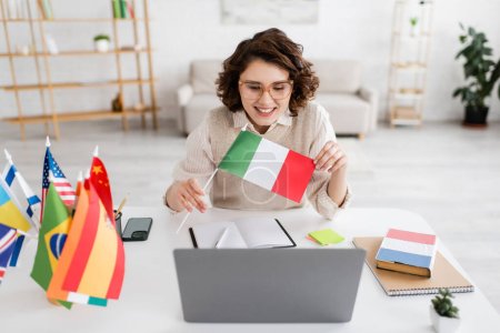 Téléchargez les photos : Professeur de langue joyeux montrant drapeau italien pendant la leçon en ligne sur ordinateur portable près des ordinateurs portables et smartphone - en image libre de droit