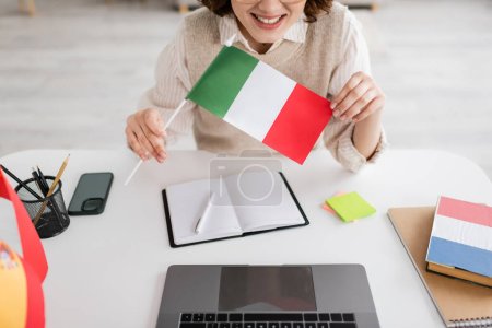 vista recortada del profesor de idiomas sonriente sosteniendo la bandera italiana cerca de dispositivos y cuadernos en la mesa en casa