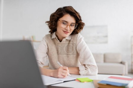 Lächelnde Frau mit Brille, die Notizen macht, während sie Fremdsprachen lernt und Online-Unterricht am Laptop anschaut 