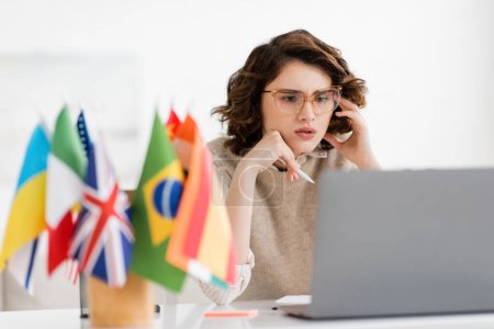 profesor de idiomas concentrado en gafas mirando el portátil cerca de banderas internacionales en primer plano borrosa 