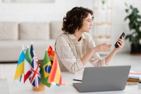 young language teacher in glasses holding smartphone near laptop and international flags on blurred foreground  Poster 645933662