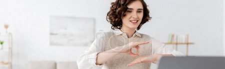 Téléchargez les photos : Professeur heureux avec les cheveux bouclés montrant geste de langue des signes pendant la leçon en ligne sur ordinateur portable, bannière - en image libre de droit