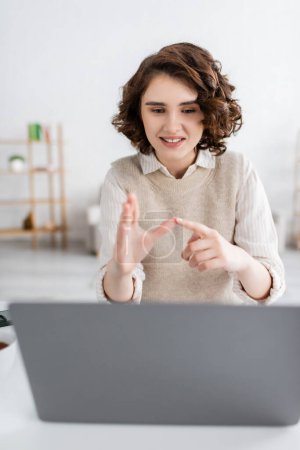 cheerful woman with curly hair teaching sign language alphabet near blurred laptop at home