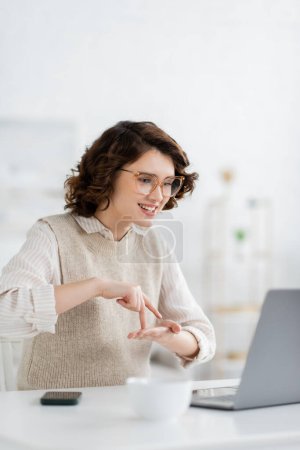 glückliche Frau zeigt Standwort, während sie Gebärdensprache während der Online-Lektion am Laptop lehrt 