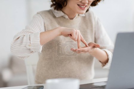 abgeschnittene Ansicht einer lächelnden Frau, die beim Unterrichten der Gebärdensprache während der Online-Lektion auf dem Laptop ein Standwort zeigt 