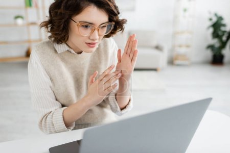 young teacher in glasses showing two handed sign language gesture while looking at laptop 