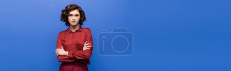 Photo for Young language teacher in burgundy blouse standing with crossed arms and looking at camera isolated on blue, banner - Royalty Free Image