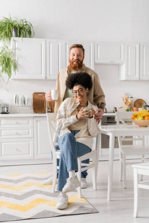Photo pour Joyeux couple multiethnique tenant des tasses à café et regardant la caméra dans la cuisine - image libre de droit