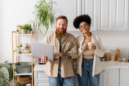Smiling interracial couple with laptop and cup of coffee looking at camera in kitchen 