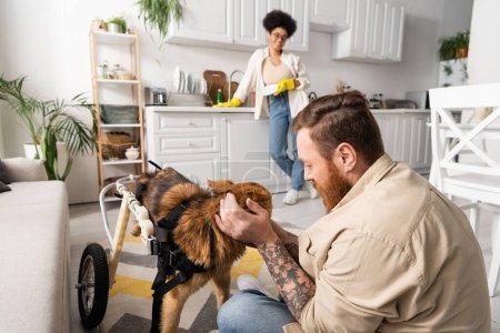 Tattooed man petting disabled dog on wheelchair near blurred african american girlfriend in kitchen 