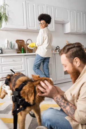 Smiling african american woman washing plate and looking at blurred boyfriend petting disabled dog at home