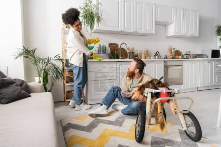 Cheerful african american woman cleaning kitchen near boyfriend and disabled dog in wheelchair at home 
