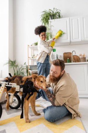 Bearded man petting disabled dog on wheelchair near african american girlfriend at home 