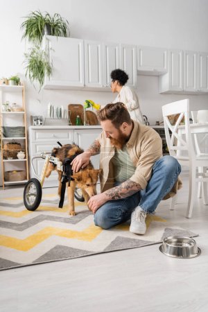 Hombre tatuado acariciando perro discapacitado en silla de ruedas cerca de cuenco en el suelo en casa 
