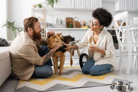 African american woman holding coffee cup near boyfriend and disabled dog in kitchen 