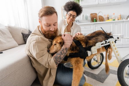 Tattooed man petting handicapped dog near african american girlfriend at home 