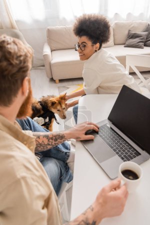 Cheerful african american woman petting handicapped dog near blurred boyfriend with laptop and coffee at home 