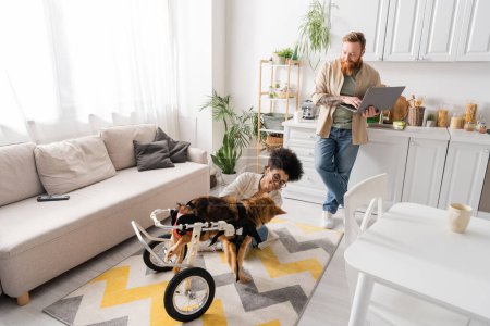 Smiling man using laptop near african american girlfriend and handicapped dog at home 